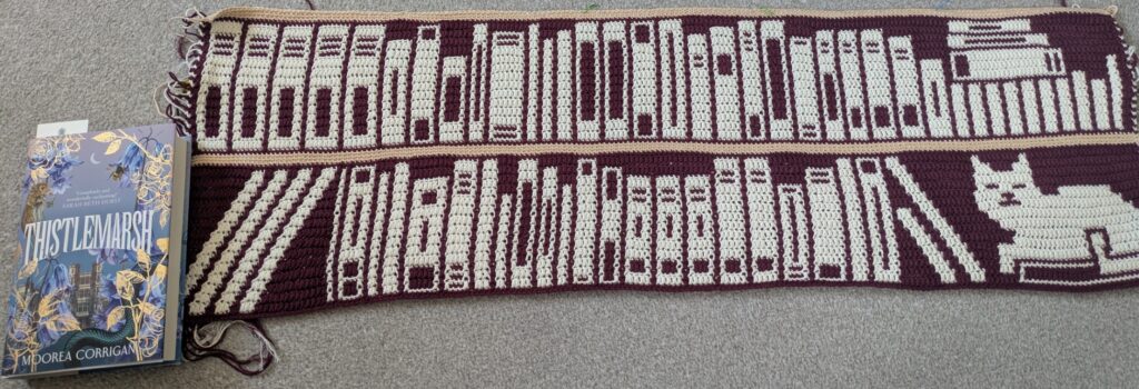 Photo of a blanket made in mosaic overlay crochet. It shows two "shelves" of a bookcase so far, mostly filled with books, with a cat sitting in the lower right on top of a book. The background colour is a deep purple-red, while the book/cat details are in cream, and the actual shelves are a tan colour. A hardback book is included for scale, showing the two shelves so far are a bit taller than it.