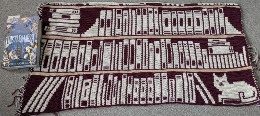 Photo of a blanket made in mosaic overlay crochet. It shows three "shelves" of a bookcase so far, mostly filled with books, with a cat sitting in the lower right on top of a book. The background colour is a deep purple-red, while the book/cat details are in cream, and the actual shelves are a tan colour. A hardback book is included for scale, about 1.5 "shelves" high.