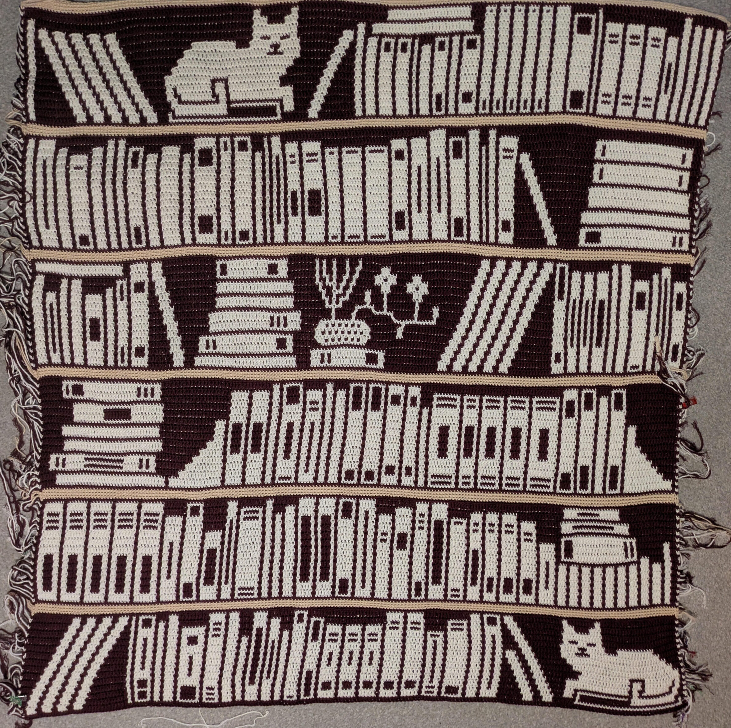 Photo of my mosaic overlay crochet blanket. It looks like a bookshelf, with six shelves so far. On the lowest shelf is a cat sat on a book, while most of the other shelves are covered in books. There's an ikebana on the fourth shelf, and another cat sat in the midst of the books on the top shelf.